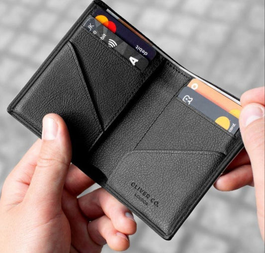 A black textured leather bifold wallet held open by two hands, showing card slots and interior pockets with demo cards inserted, against a blurred gray outdoor background.