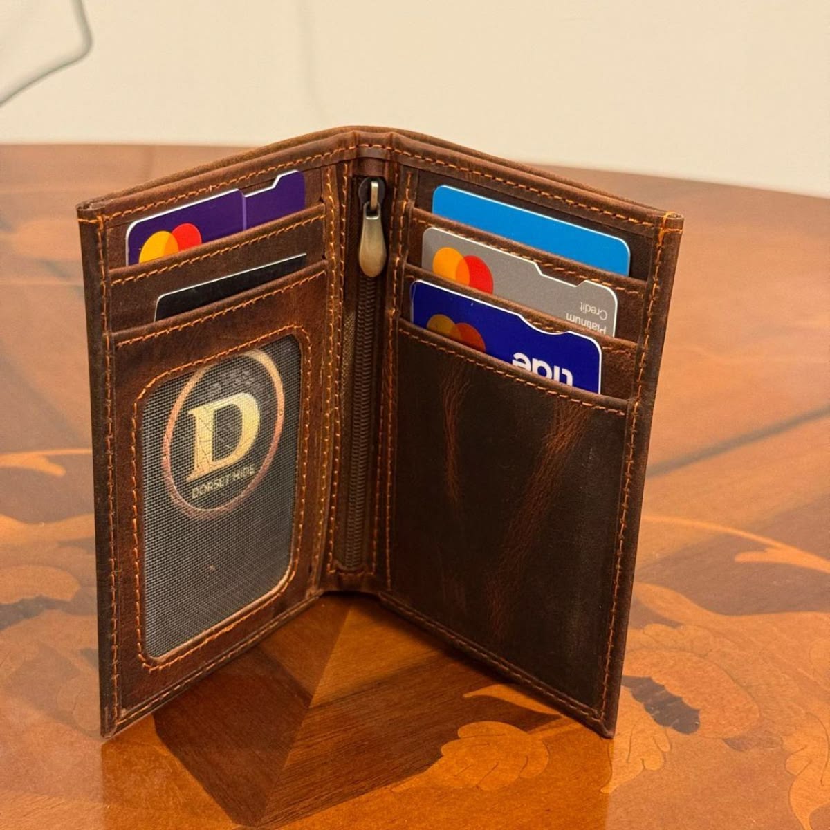 An open brown leather bifold wallet standing on a wooden table, showing card slots, a zipper, and a circular ID window with a logo.