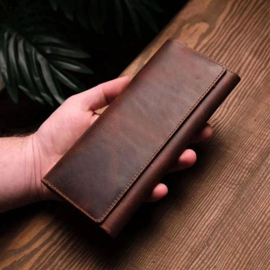 A hand holding a long brown leather wallet or clutch with a fold-over flap, shown against a wooden tabletop background with dark green leaves in the upper left.