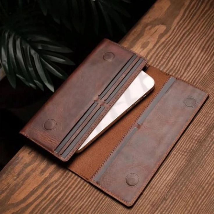 A brown leather phone wallet case with multiple card slots and a phone partially inserted, photographed on a wooden tabletop with dark green leaves in the background.