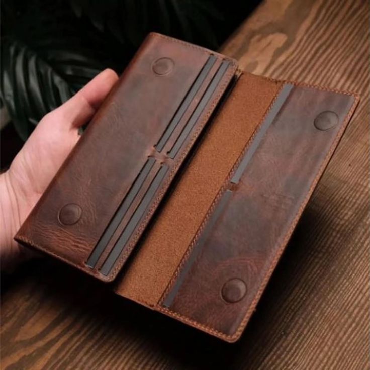 A hand holds an open brown leather wallet or card holder with multiple card slots and snap-button details, set against a dark wood surface.